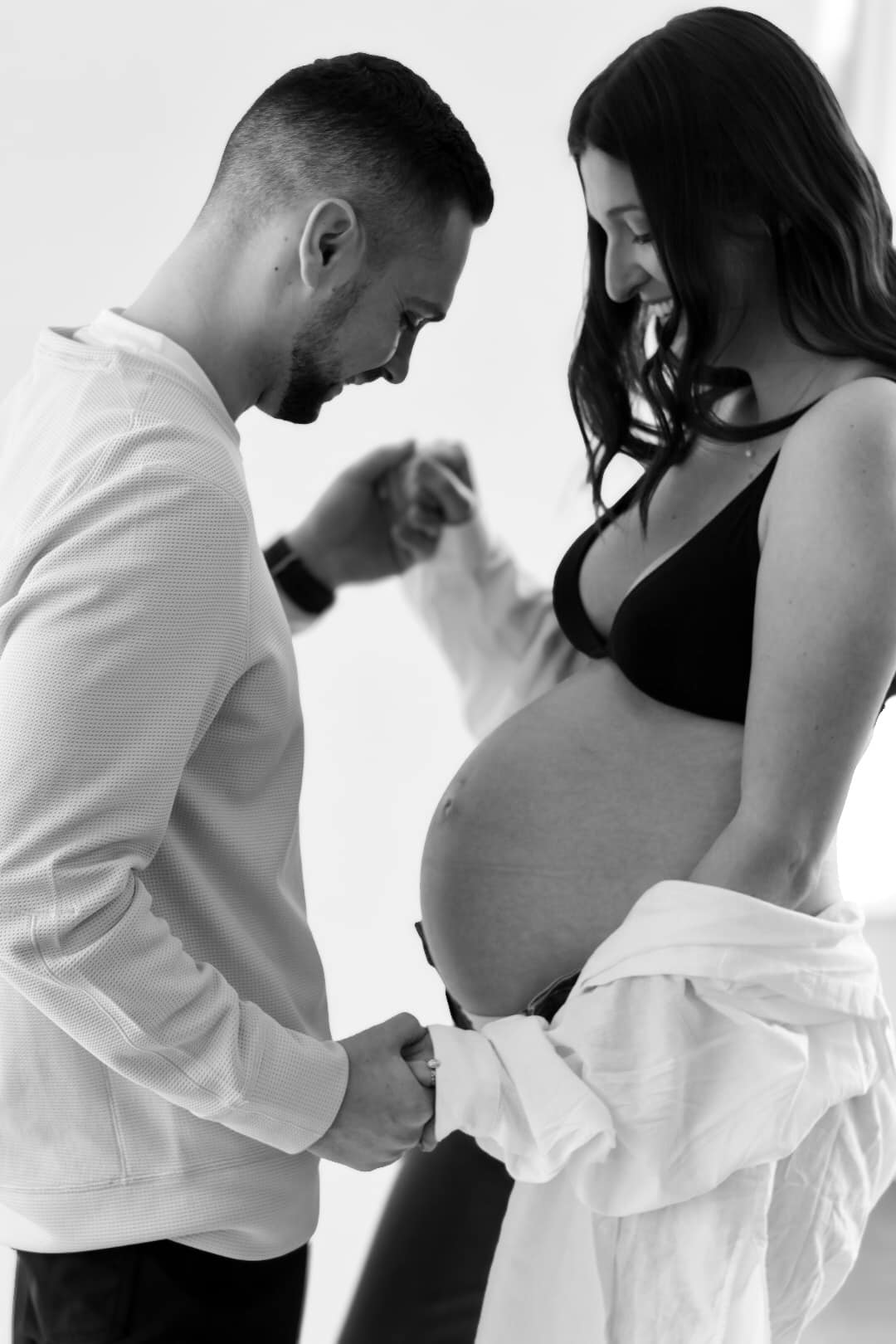 Black and white editorial maternity portrait, expectant couple holding hands beside the bump