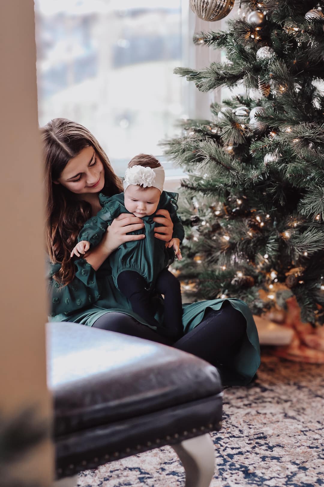Older sister in green holiday dress holding her baby sister by a Christmas tree