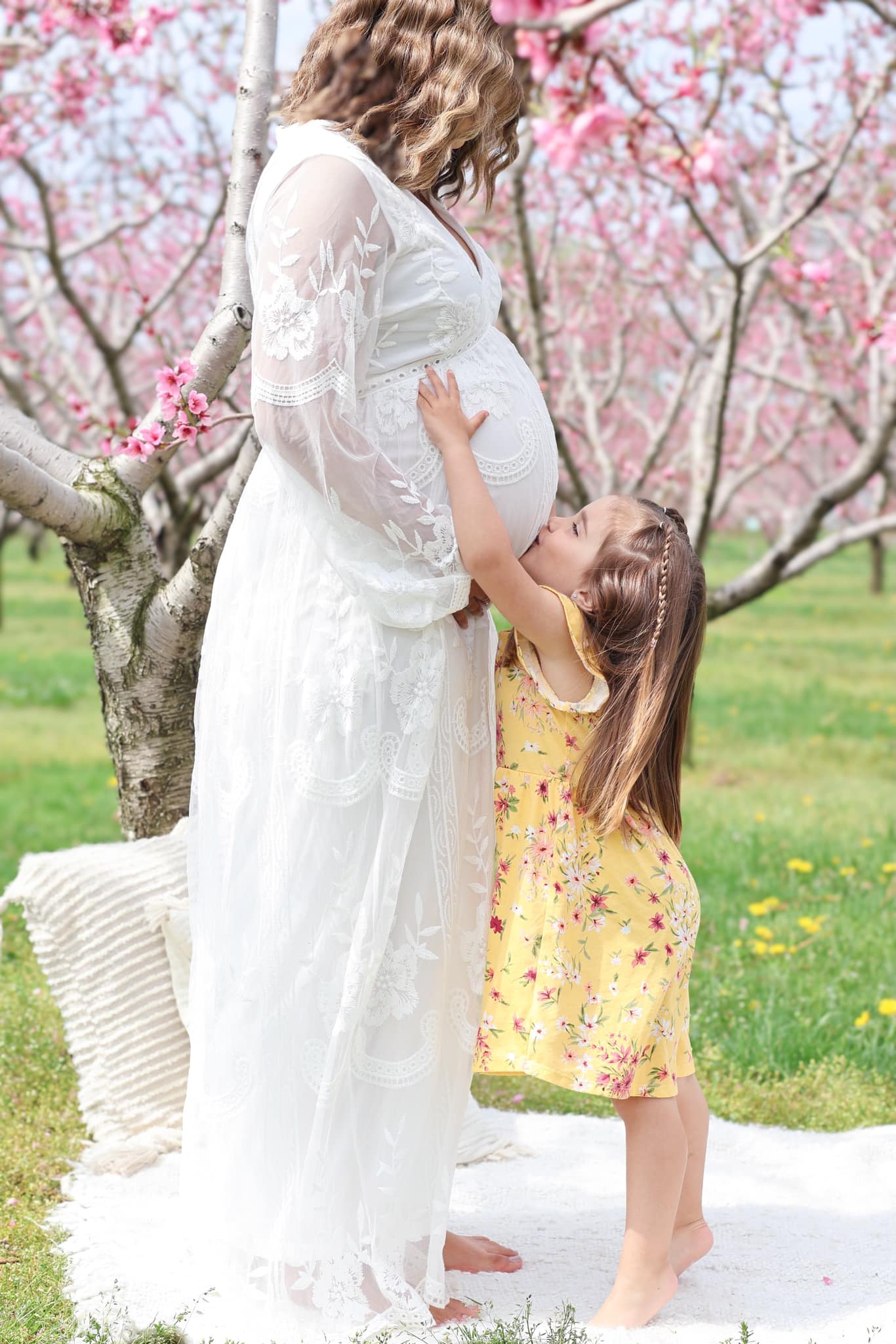Older daughter in yellow dress kissing her pregnant mother’s belly in a peach blossom orchard